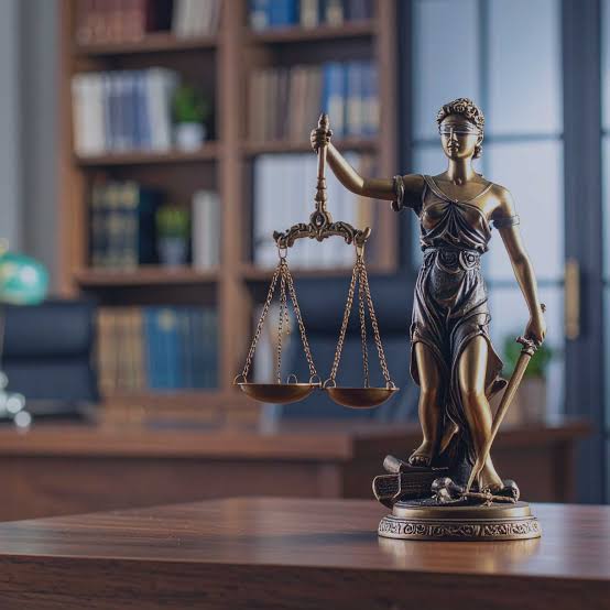 Estátua da Justiça com balança em destaque, dentro de um escritório jurídico com biblioteca, representando a administração judicial