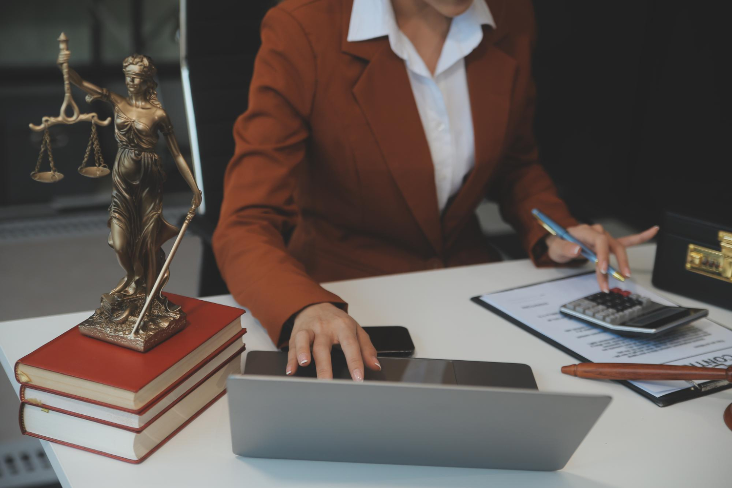 Profissional em escritório de advocacia com laptop, calculadora, livros jurídicos e estátua da Justiça, representando a contabilidade especializada para advogados